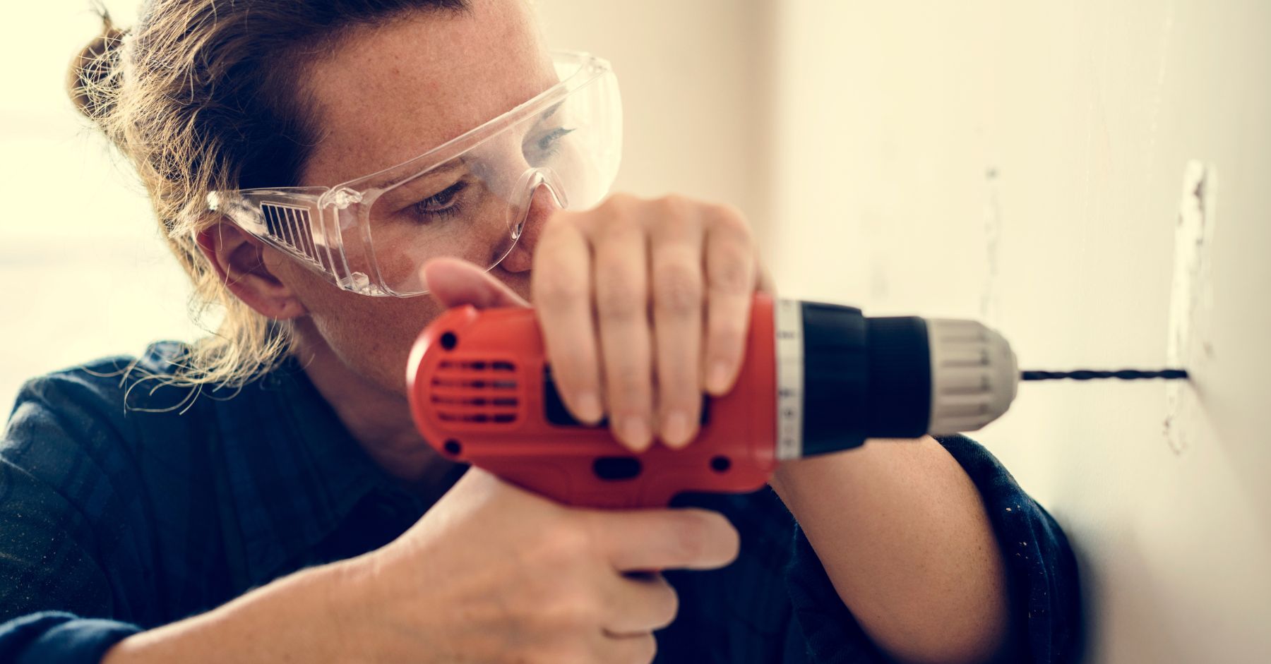 1.5 million homes target: A woman wearing clear safety goggles and a dark shirt using a red power drill to bore a hole into a white wall.
