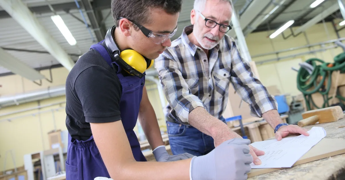 An older carpenter teaching a younger apprentice