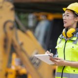 A young woman on a construction site in a hi vis, hard hat, and ear protectors uses a clipboard