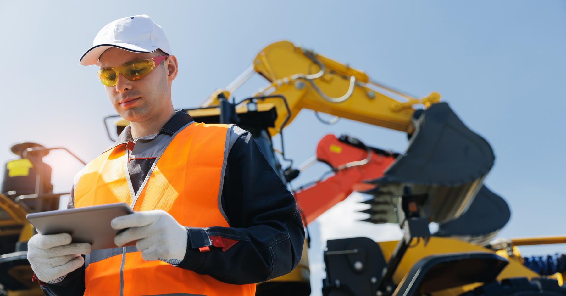 Construction certifications that help you get hired A construction worker wearing a hi-vis vest, hard hat and safety glasses checks a tablet, with an excavator visible in the background.