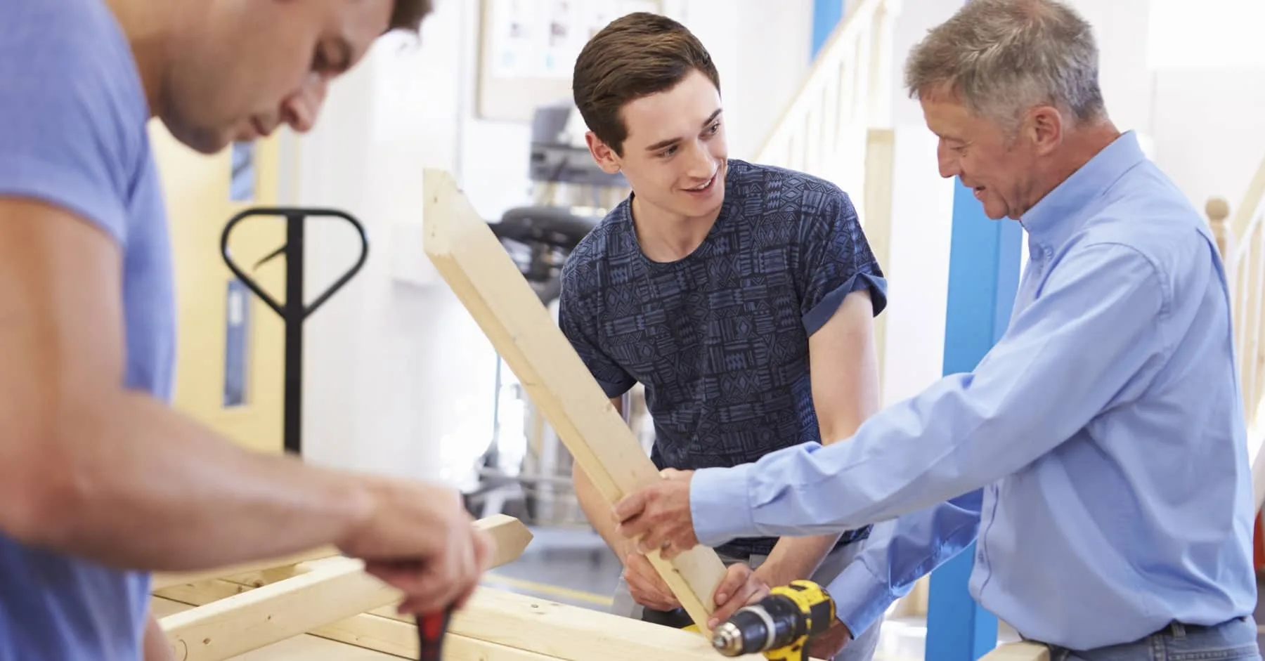 Older tradesman instructing a young man in carpentry, holding a wooden beam, while another worker uses a tool in the foreground
