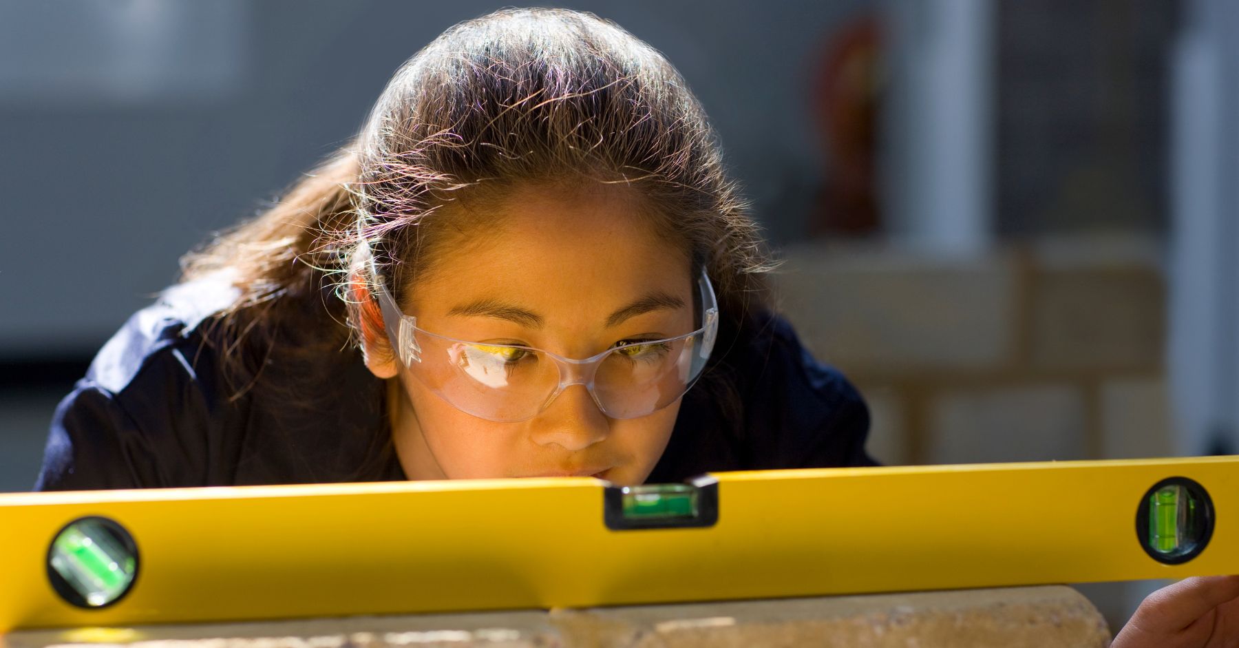 How to Get Into Construction With No Experience: Young woman in safety glasses closely checking a yellow spirit level resting on a brick.