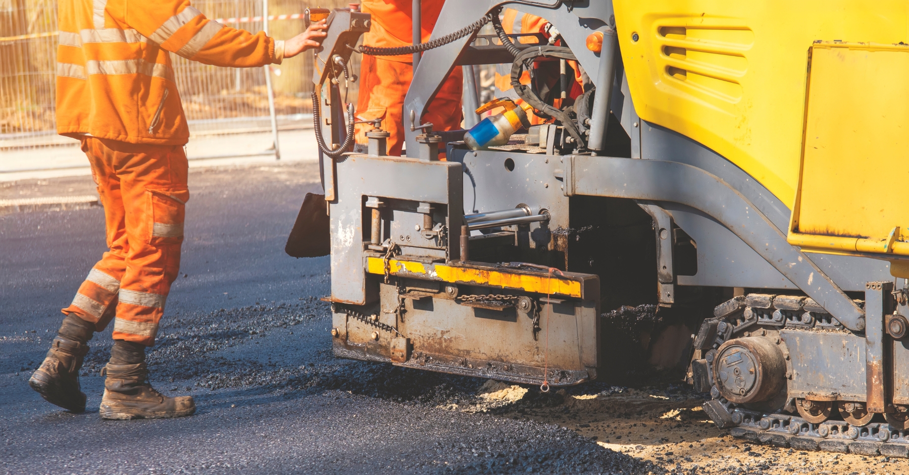 Two road workers wearing orange high-visibility clothing operate heavy machinery on a newly laid tarmac surface. The image captures close-up detail of the paving equipment and work boots on a busy construction site.