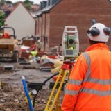 Most in-demand construction skills A construction worker in a hi-vis jacket and hard hat viewed from behind, operating a surveying instrument on a tripod at a building site.