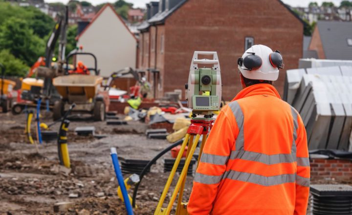 Most in-demand construction skills A construction worker in a hi-vis jacket and hard hat viewed from behind, operating a surveying instrument on a tripod at a building site.