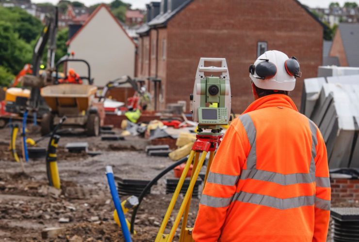 Most in-demand construction skills A construction worker in a hi-vis jacket and hard hat viewed from behind, operating a surveying instrument on a tripod at a building site.