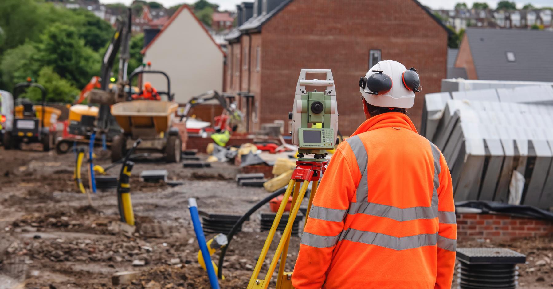 Most in-demand construction skills A construction worker in a hi-vis jacket and hard hat viewed from behind, operating a surveying instrument on a tripod at a building site.