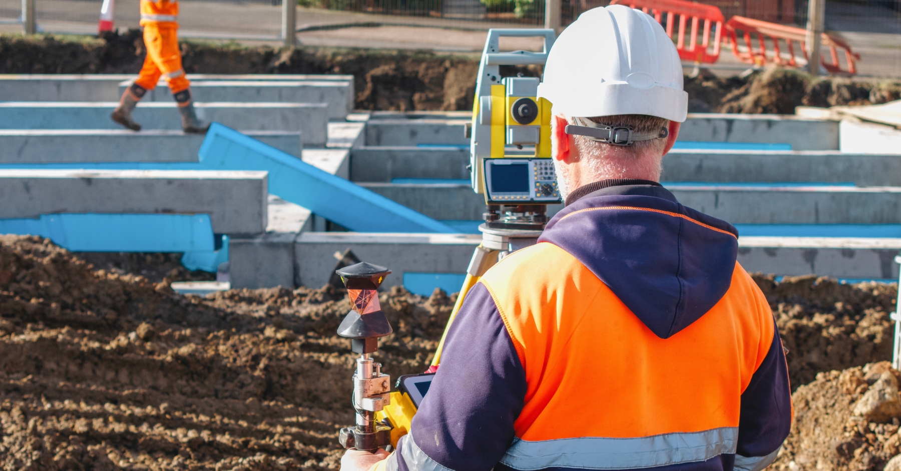 A construction worker wearing a hard hat and high-visibility jacket operates a surveying instrument on a building site. In the background, another worker moves among concrete foundations and blue steel structures under bright daylight.