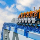 A rollercoaster filled with passengers raises its orange seats into the sky, with riders lifting their hands in excitement. The blue steel track and bright sky in the background convey motion and thrill at a theme park.