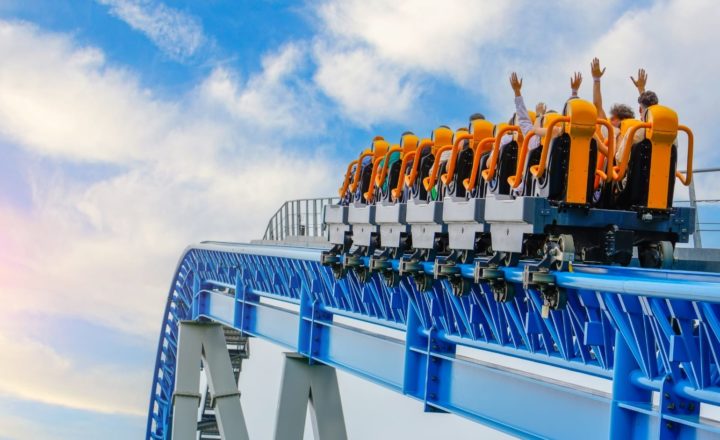 A rollercoaster filled with passengers raises its orange seats into the sky, with riders lifting their hands in excitement. The blue steel track and bright sky in the background convey motion and thrill at a theme park.