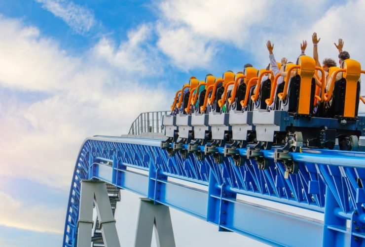 A rollercoaster filled with passengers raises its orange seats into the sky, with riders lifting their hands in excitement. The blue steel track and bright sky in the background convey motion and thrill at a theme park.