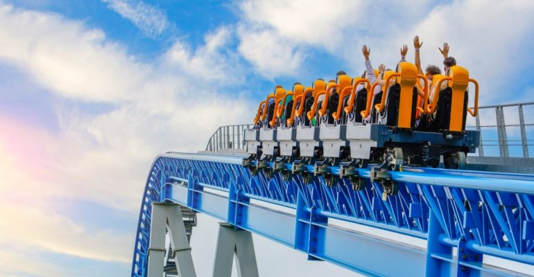 A rollercoaster filled with passengers raises its orange seats into the sky, with riders lifting their hands in excitement. The blue steel track and bright sky in the background convey motion and thrill at a theme park.