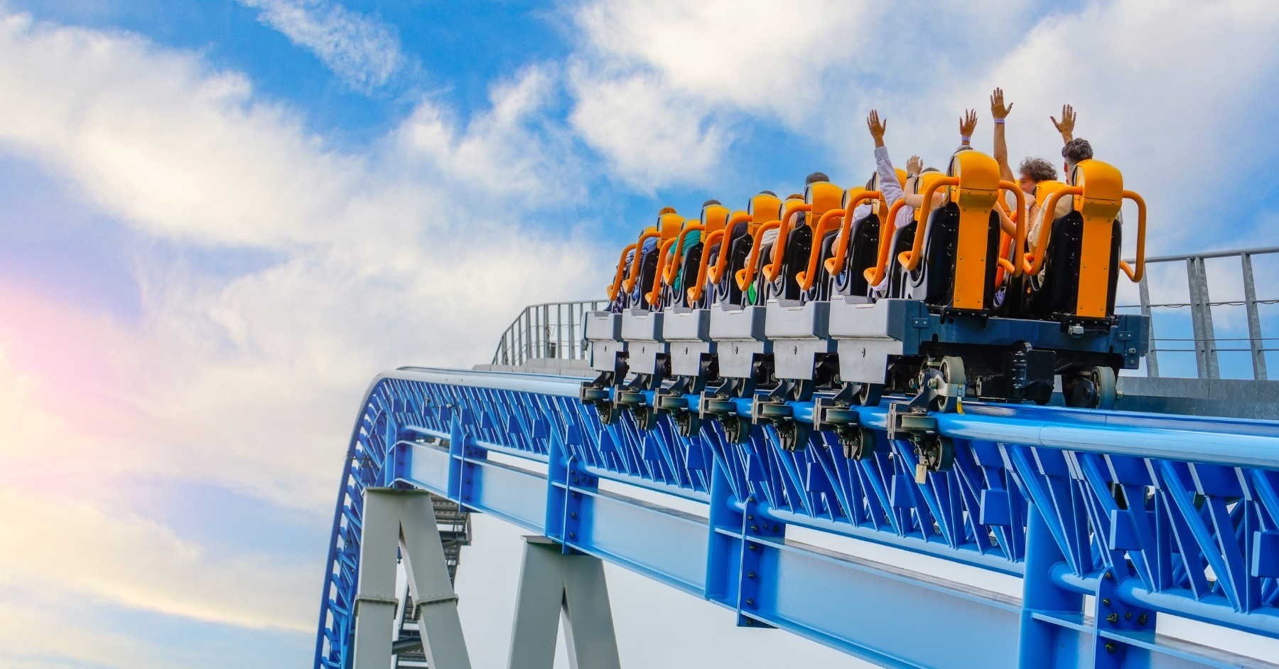 A rollercoaster filled with passengers raises its orange seats into the sky, with riders lifting their hands in excitement. The blue steel track and bright sky in the background convey motion and thrill at a theme park.