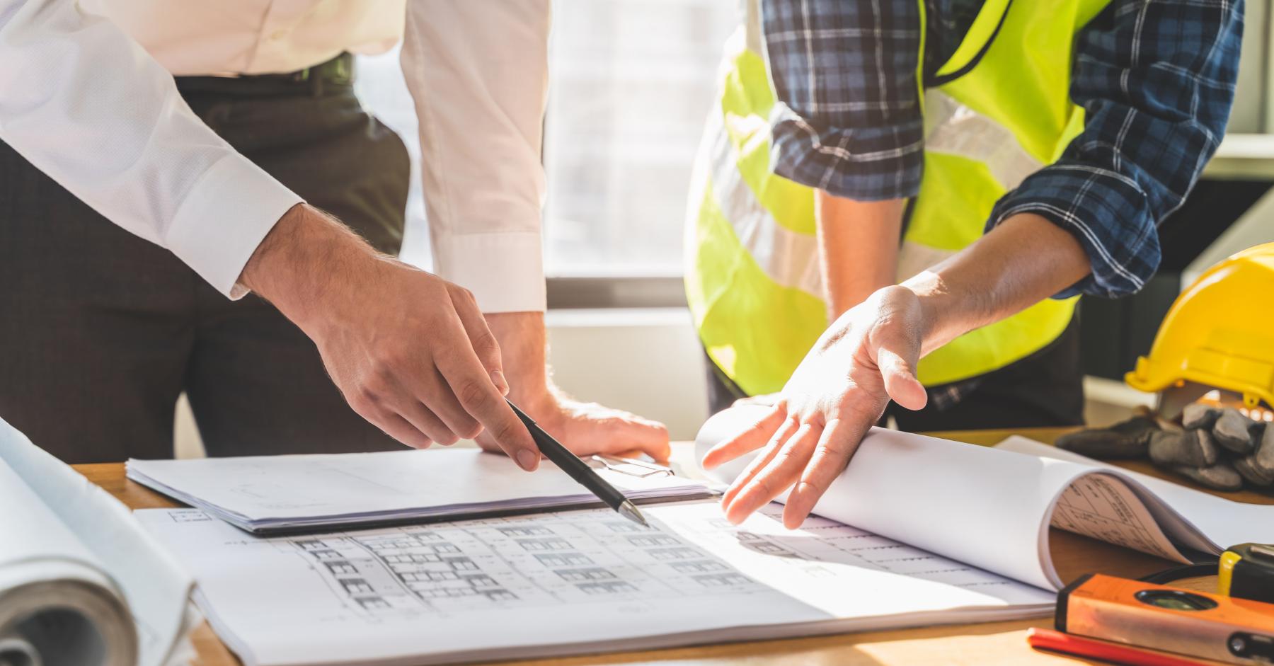 What skills do construction employers want Close-up of two people reviewing building blueprints on a table, one in business attire and one wearing a hi-vis vest, with a hard hat and tape measure nearby.