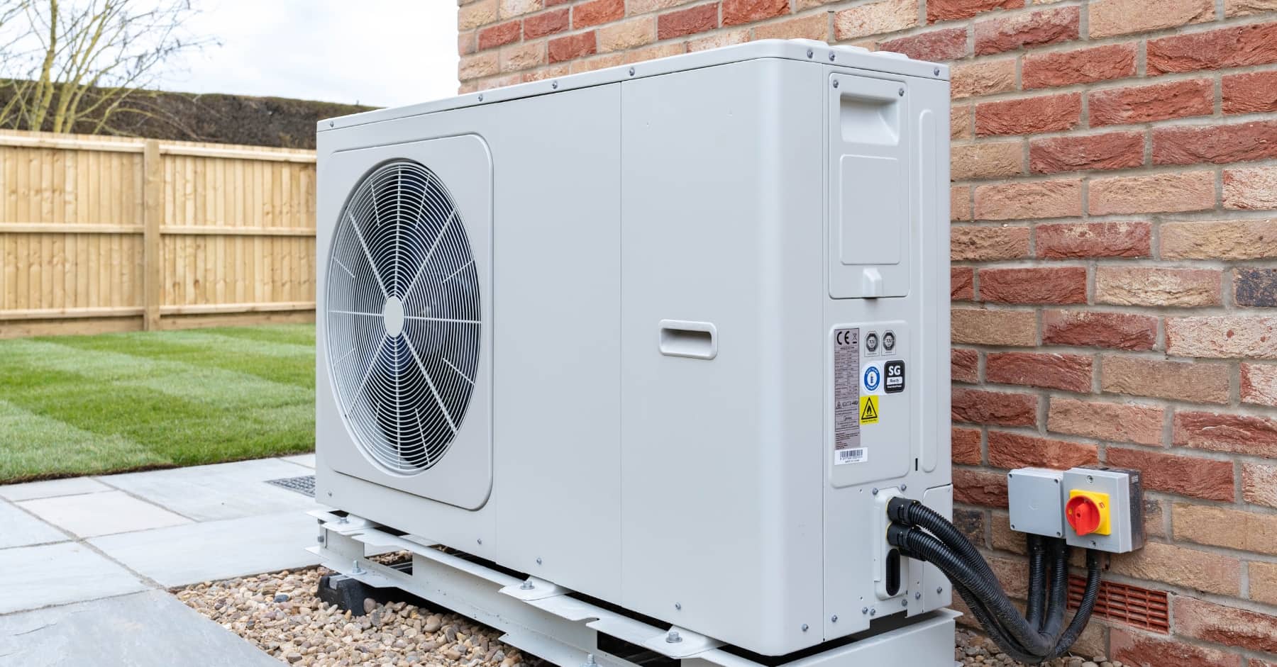 Outdoor air source heat pump unit installed beside a brick house, connected to power supply, with a fenced garden and lawn in the background.