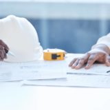 Close-up of hands working over building plans with a white safety helmet, yellow tape measure, and pencil on the table.