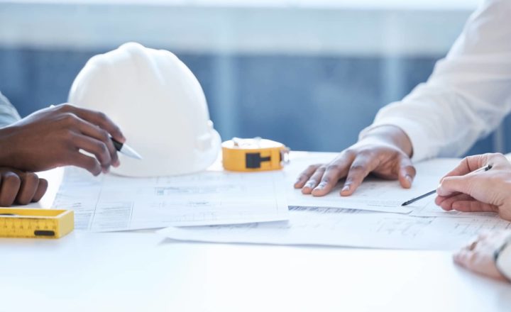 Close-up of hands working over building plans with a white safety helmet, yellow tape measure, and pencil on the table.
