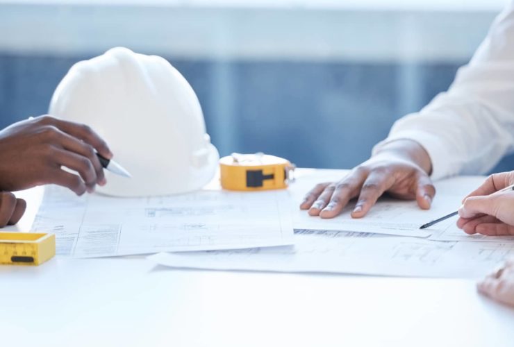 Close-up of hands working over building plans with a white safety helmet, yellow tape measure, and pencil on the table.