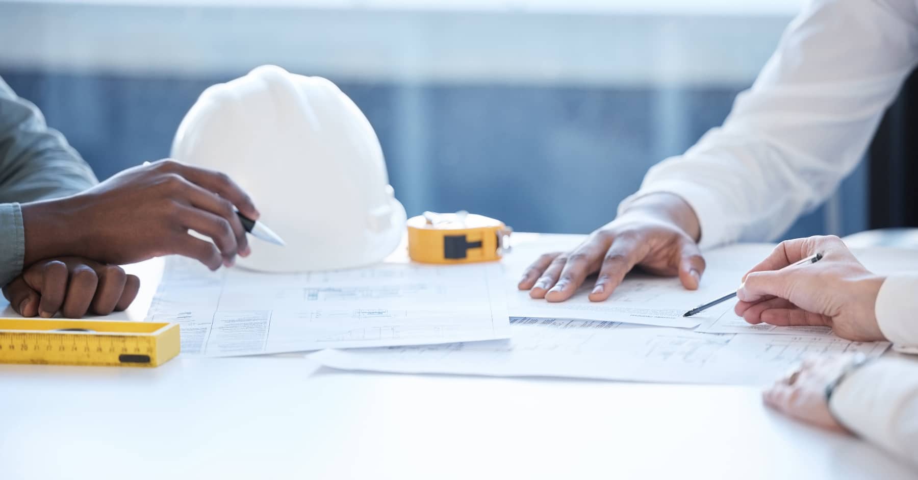 Close-up of hands working over building plans with a white safety helmet, yellow tape measure, and pencil on the table.