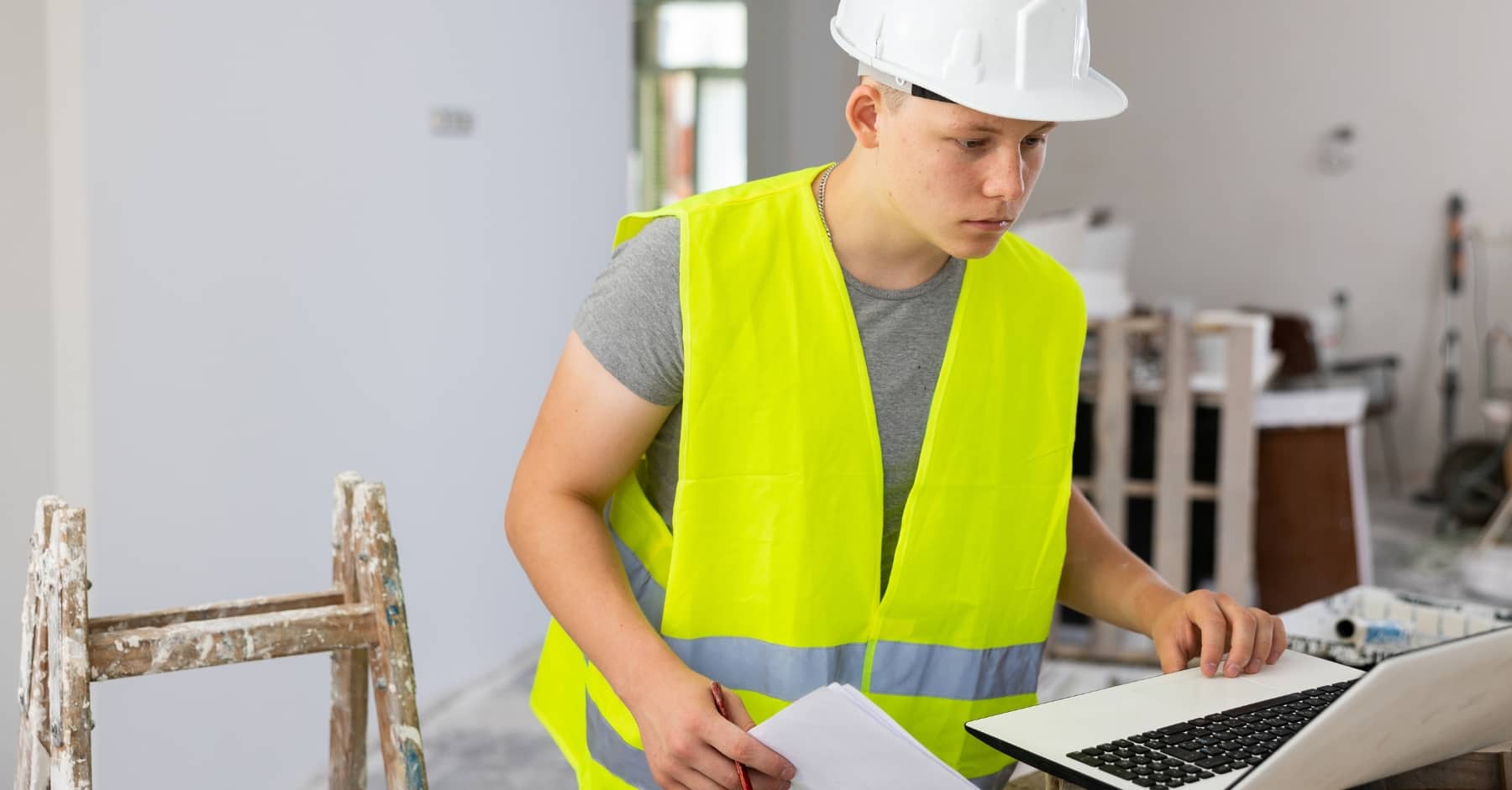 Young construction worker in a hi-vis vest and white hard hat using a laptop on-site, holding documents, with a paint-splattered ladder nearby