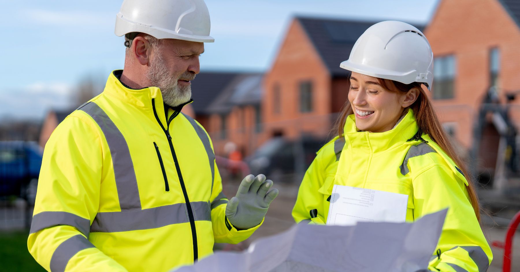 construction labour shortage UK: An older bearded man and a smiling younger woman, both in yellow hi-vis jackets and white hard hats, reviewing paperwork together in front of newly built brick houses.