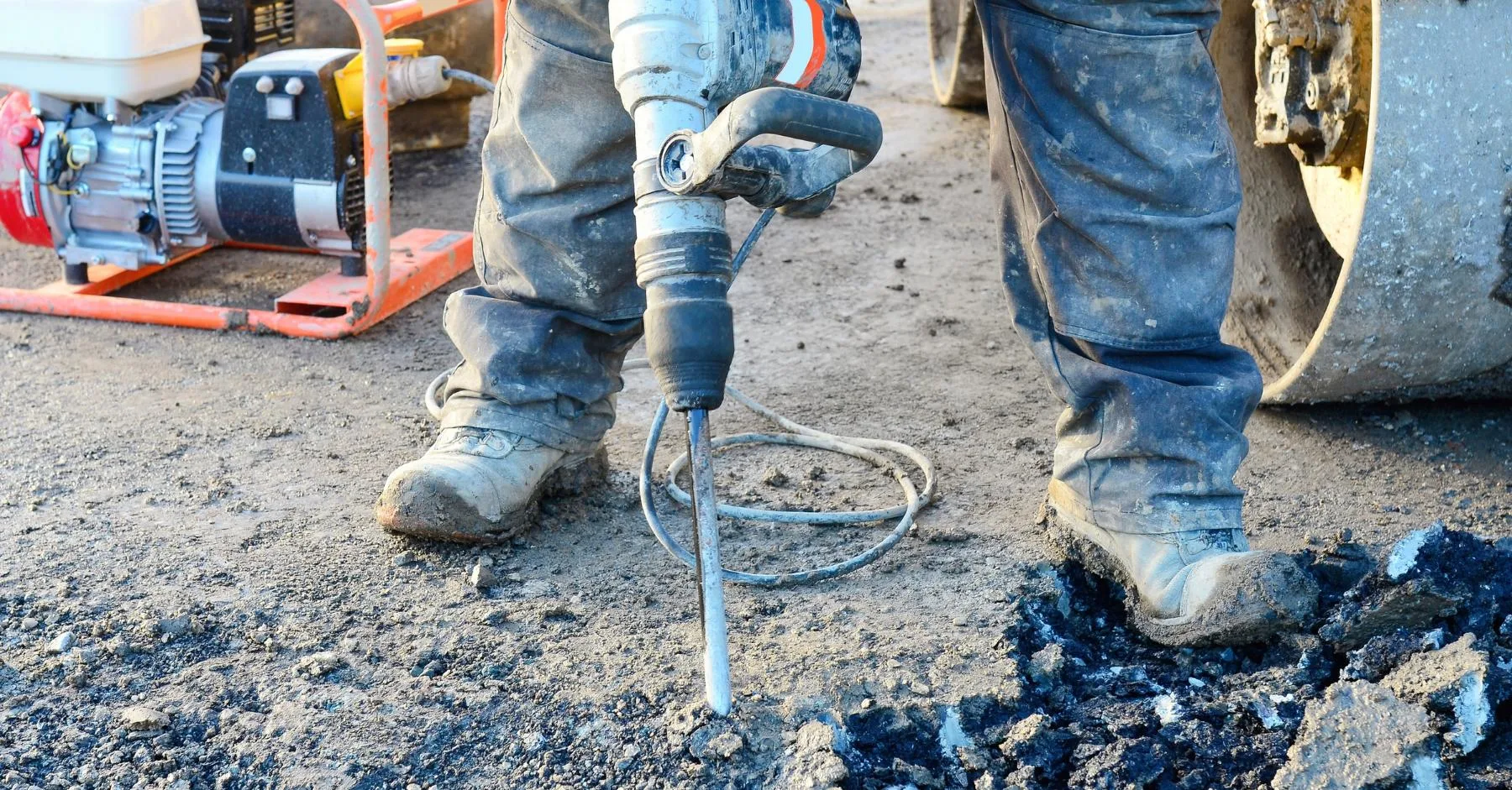 construction pay rates April 2026 UK: Close-up of a construction worker's boots and legs as they operate a jackhammer breaking up asphalt on a worksite
