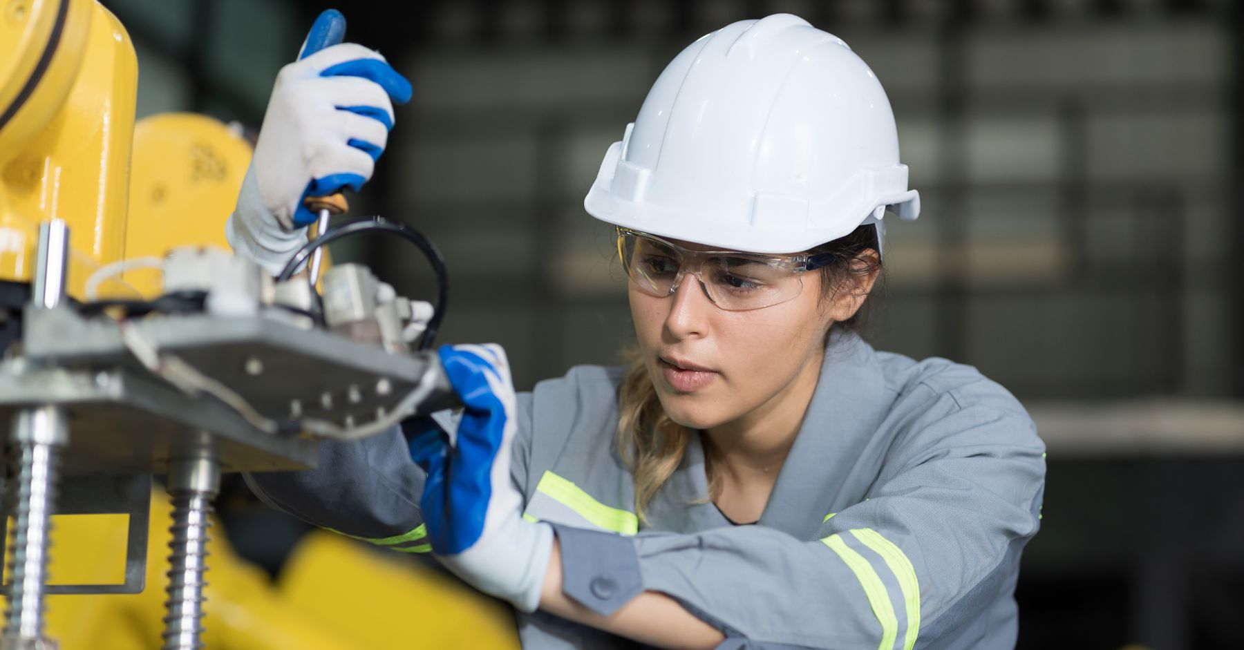 construction recruitment agency budget 2026 impact: A female engineer wearing a white hard hat, safety glasses, blue work gloves and grey hi-vis overalls using a screwdriver to work on yellow industrial machinery.