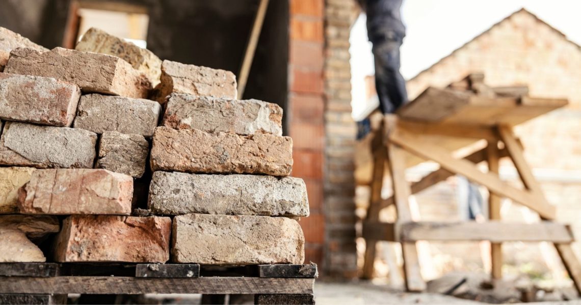 construction skills shortage: A stack of weathered reclaimed bricks on a wooden pallet in the foreground, with a blurred worker standing on a raised platform beside a brick building in the background.