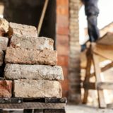 construction skills shortage: A stack of weathered reclaimed bricks on a wooden pallet in the foreground, with a blurred worker standing on a raised platform beside a brick building in the background.