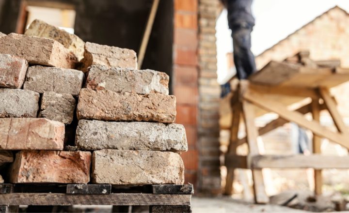 construction skills shortage: A stack of weathered reclaimed bricks on a wooden pallet in the foreground, with a blurred worker standing on a raised platform beside a brick building in the background.