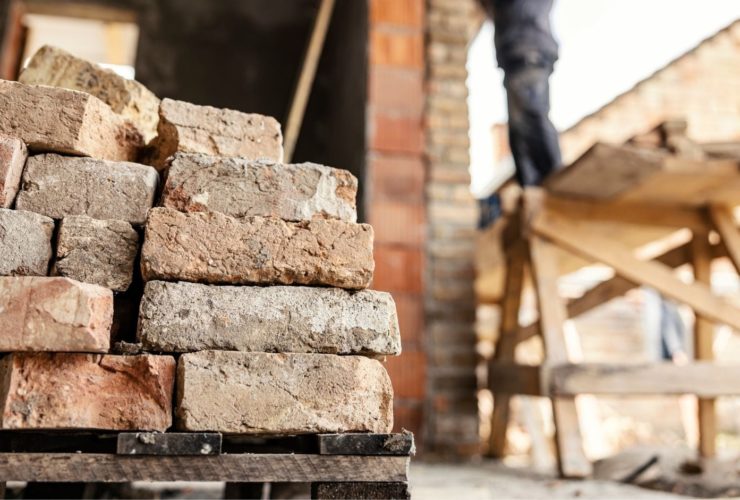 construction skills shortage: A stack of weathered reclaimed bricks on a wooden pallet in the foreground, with a blurred worker standing on a raised platform beside a brick building in the background.
