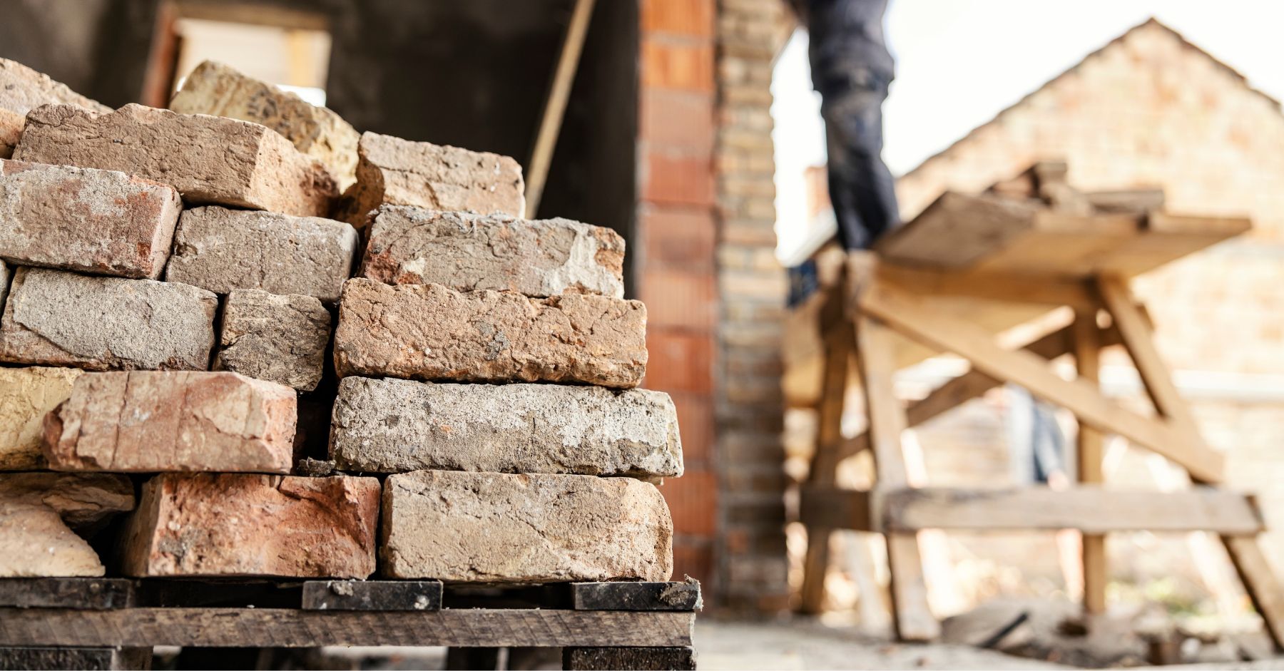 construction skills shortage: A stack of weathered reclaimed bricks on a wooden pallet in the foreground, with a blurred worker standing on a raised platform beside a brick building in the background.