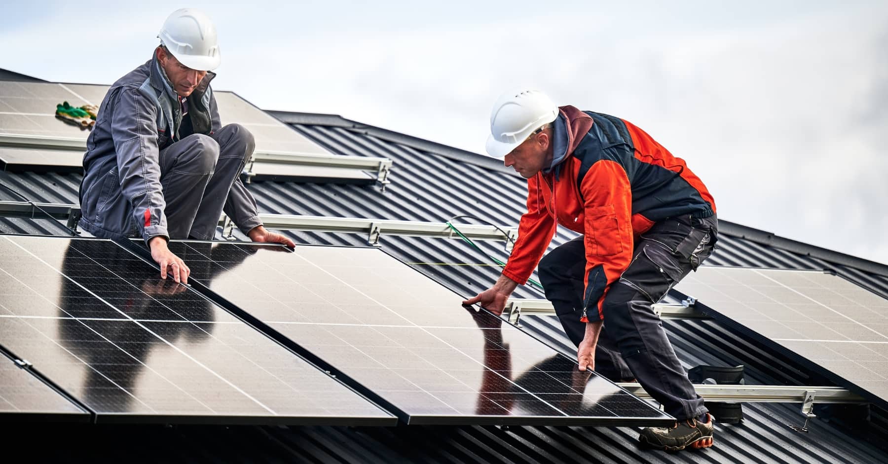 Two construction workers in safety gear installing solar panels on a sloped roof under a cloudy sky
