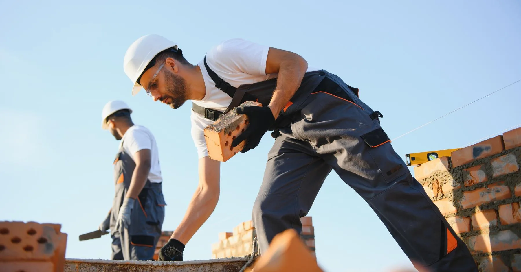 how April 2026 wage changes affect construction: Two construction workers in hard hats and dungarees laying bricks on a wall against a clear blue sky