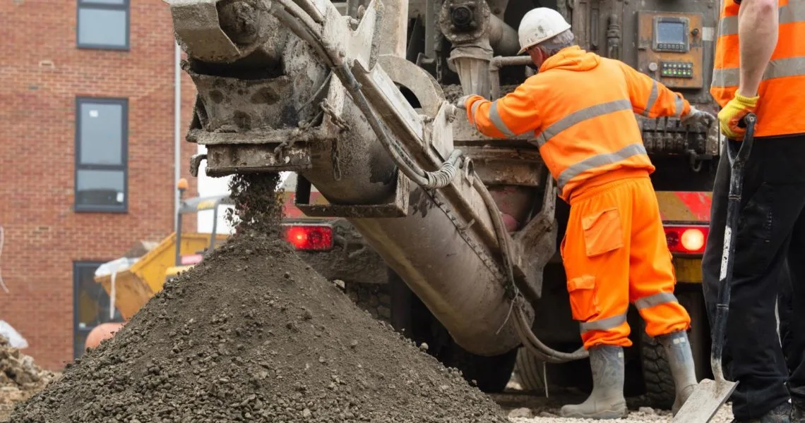industrial construction career opportunities UK A construction worker in an orange high-visibility suit and white hard hat operates heavy machinery on a building site, with wet concrete or aggregate being discharged from the equipment into a large pile, and a brick building visible in the background.