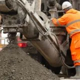 industrial construction career opportunities UK A construction worker in an orange high-visibility suit and white hard hat operates heavy machinery on a building site, with wet concrete or aggregate being discharged from the equipment into a large pile, and a brick building visible in the background.