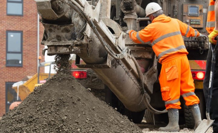 industrial construction career opportunities UK A construction worker in an orange high-visibility suit and white hard hat operates heavy machinery on a building site, with wet concrete or aggregate being discharged from the equipment into a large pile, and a brick building visible in the background.
