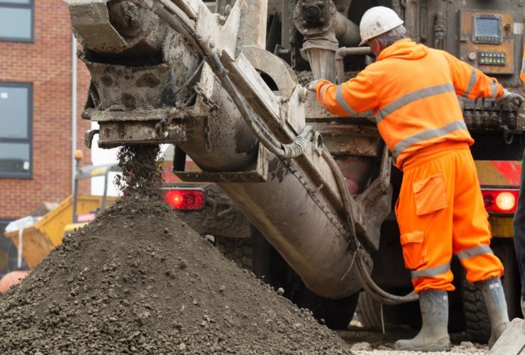 industrial construction career opportunities UK A construction worker in an orange high-visibility suit and white hard hat operates heavy machinery on a building site, with wet concrete or aggregate being discharged from the equipment into a large pile, and a brick building visible in the background.