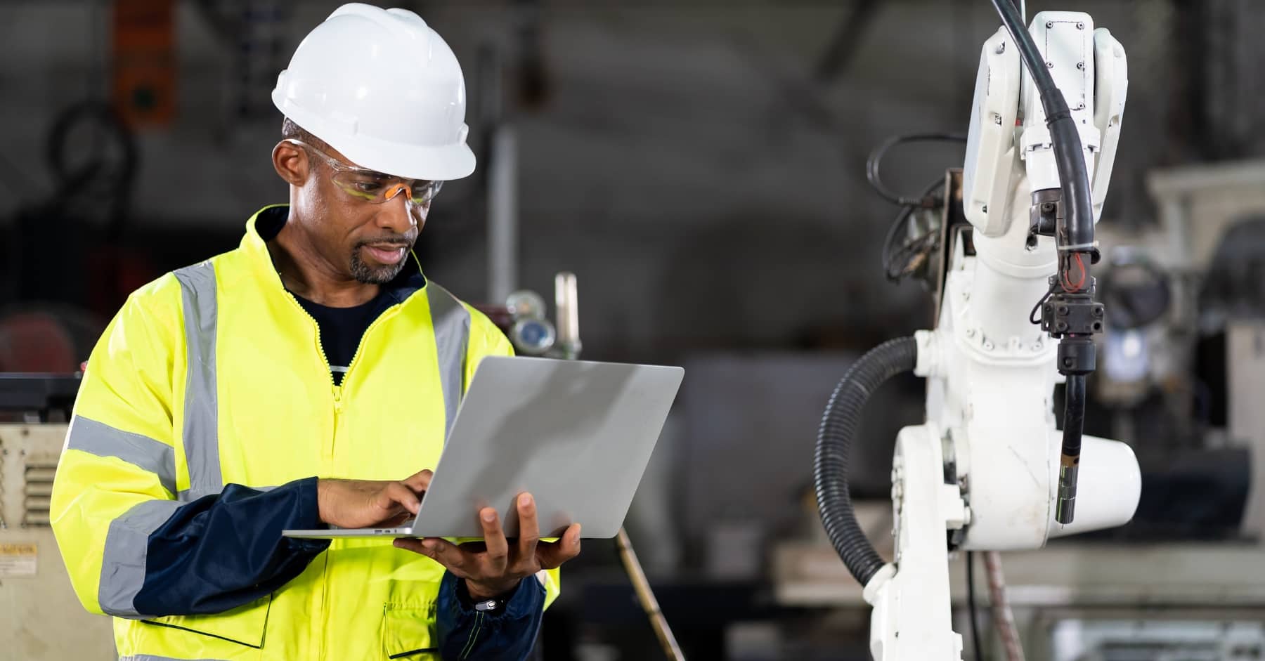 Construction worker in hi-vis jacket and white hard hat using a laptop beside industrial machinery, wearing safety goggles