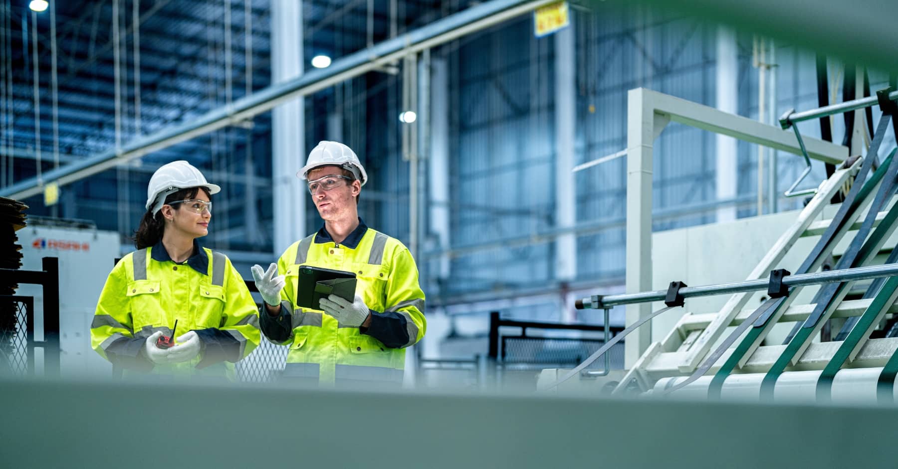 Two construction professionals in hi-vis jackets and hard hats discussing work inside a modern industrial facility, one holding a tablet and the other a radio