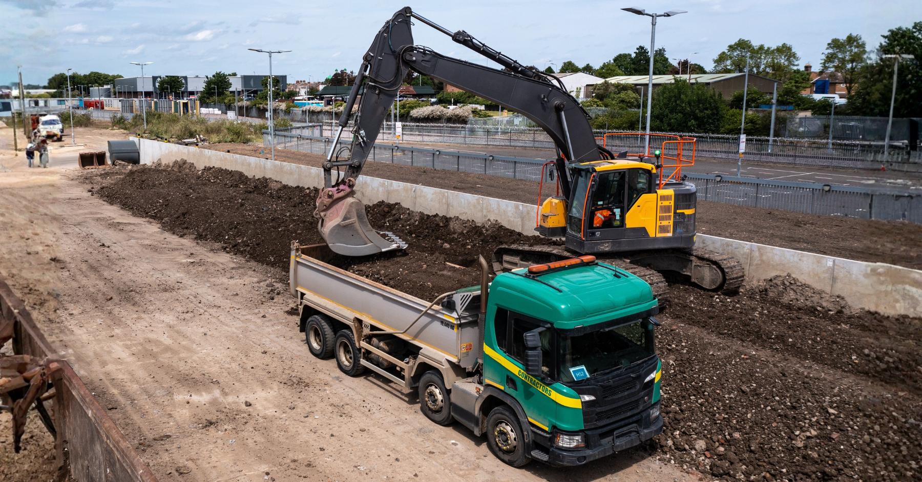 logistics and warehouse construction jobs growth UK An aerial view of a large commercial construction site, with a yellow excavator loading soil into a green tipper lorry, surrounded by cleared ground, concrete barriers and industrial buildings in the background.