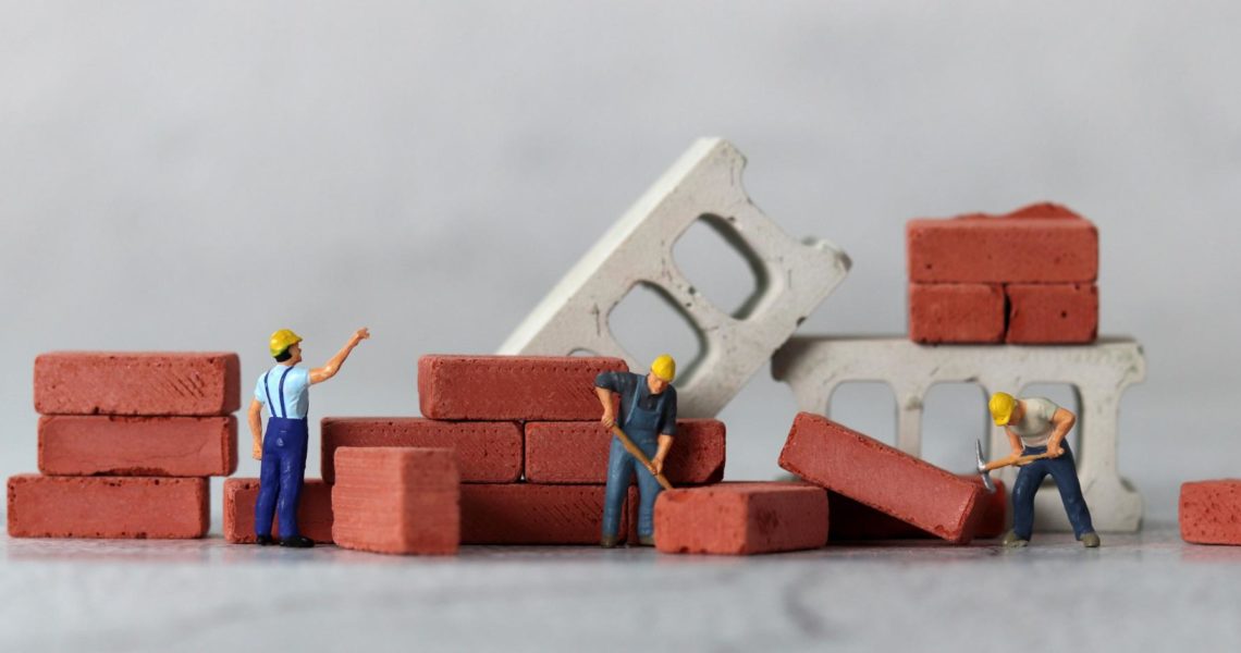 minimum wage increase 2026 construction: Miniature figurine construction workers in yellow hard hats building a small brick wall, surrounded by stacked red bricks and concrete blocks