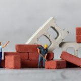 minimum wage increase 2026 construction: Miniature figurine construction workers in yellow hard hats building a small brick wall, surrounded by stacked red bricks and concrete blocks