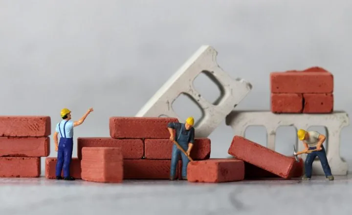 minimum wage increase 2026 construction: Miniature figurine construction workers in yellow hard hats building a small brick wall, surrounded by stacked red bricks and concrete blocks