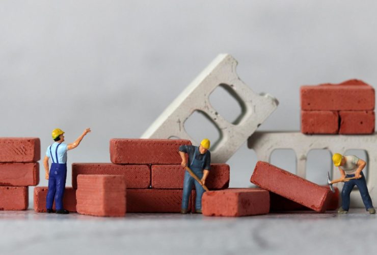 minimum wage increase 2026 construction: Miniature figurine construction workers in yellow hard hats building a small brick wall, surrounded by stacked red bricks and concrete blocks