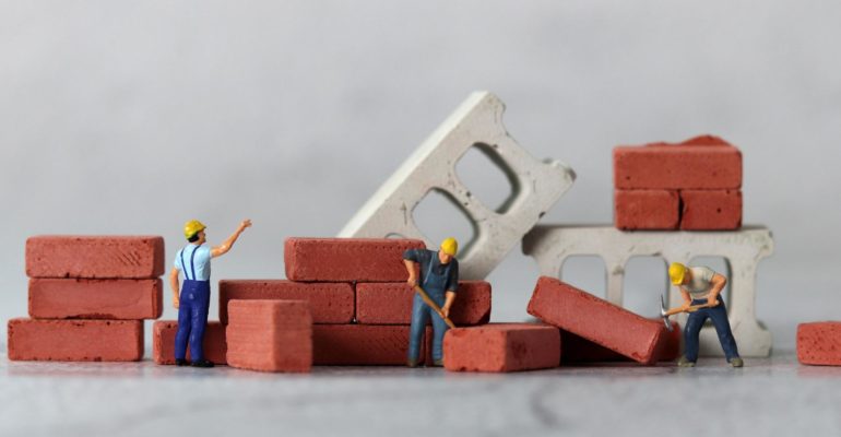 minimum wage increase 2026 construction: Miniature figurine construction workers in yellow hard hats building a small brick wall, surrounded by stacked red bricks and concrete blocks