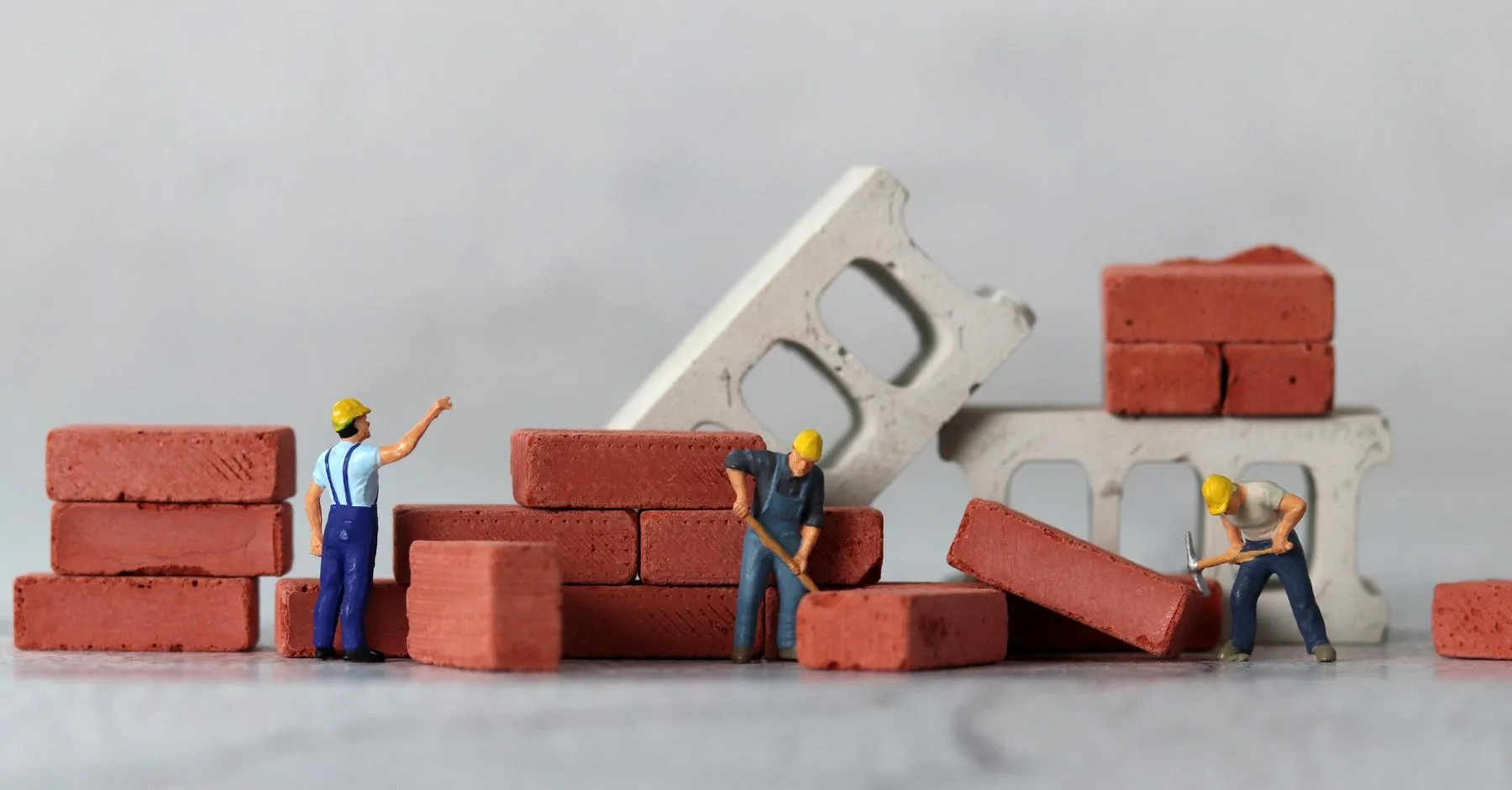 minimum wage increase 2026 construction: Miniature figurine construction workers in yellow hard hats building a small brick wall, surrounded by stacked red bricks and concrete blocks