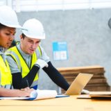 Two construction workers in hi-vis vests and hard hats reviewing documents and working on a laptop in a warehouse setting