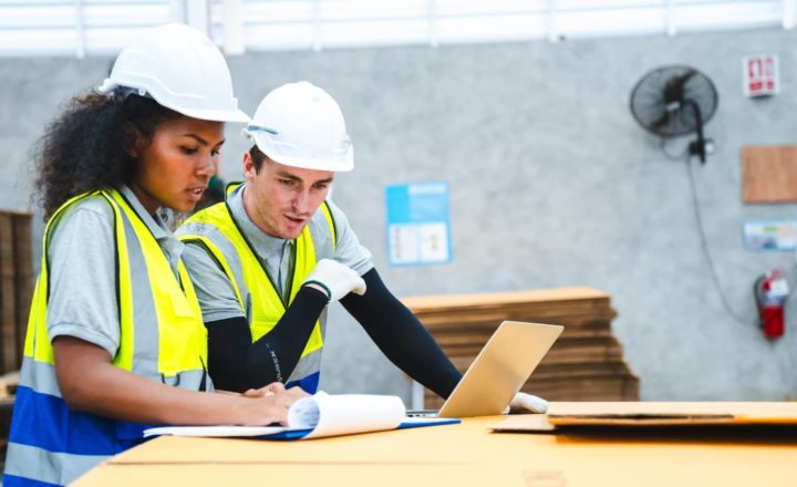 Two construction workers in hi-vis vests and hard hats reviewing documents and working on a laptop in a warehouse setting