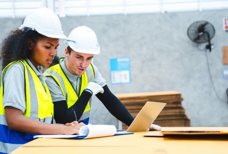 Two construction workers in hi-vis vests and hard hats reviewing documents and working on a laptop in a warehouse setting